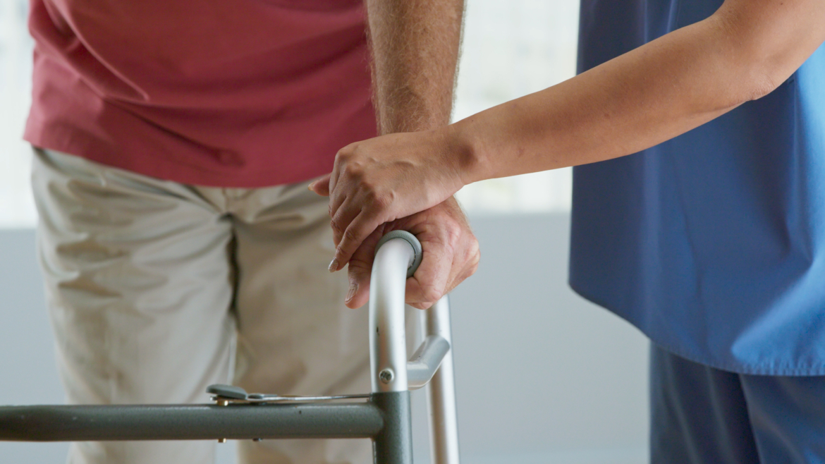 Close up on hands of physical therapist or doctor helping older male patient learn to walk with a walker after an accident.