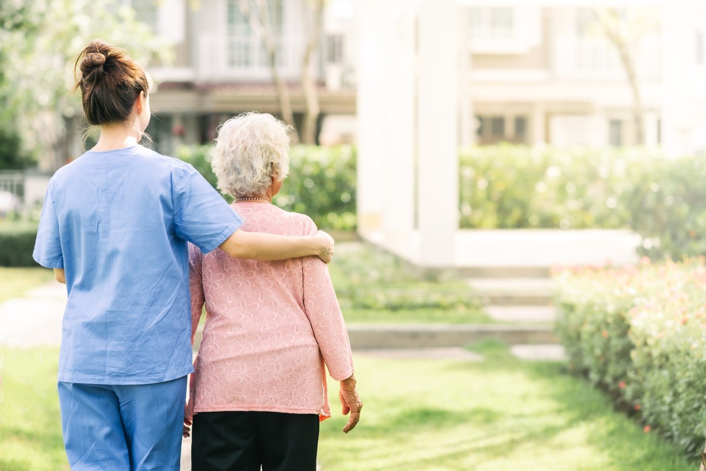 A young woman caregiver in blue scrubs places her arm around an elderly woman as they walk together outside surrounded by greenery.
