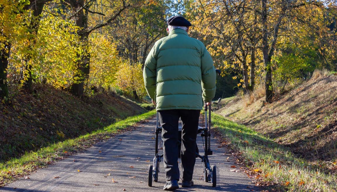 A senior man with a green winter coat walks down a dirt pathway with a rollator.
