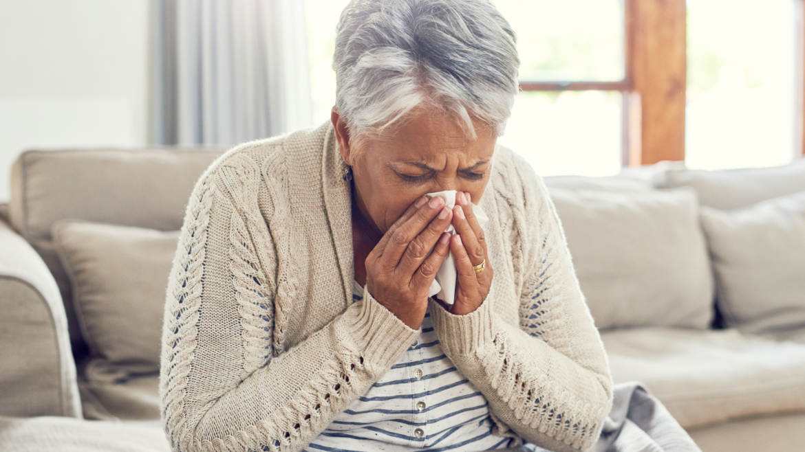 Senior woman, sick during flu season, and blowing nose at home from virus, allergy and illness on couch. Tired, tissue and elderly female person in a house lounge with sneeze from covid and flu on a sofa with allergies.