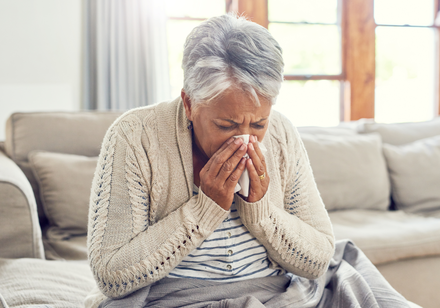 Senior woman, sick during flu season, and blowing nose at home from virus, allergy and illness on couch. Tired, tissue and elderly female person in a house lounge with sneeze from covid and flu on a sofa with allergies.