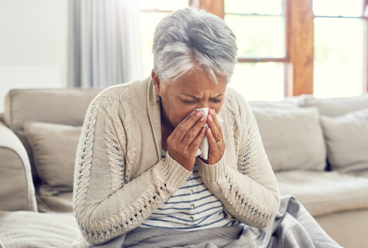 Senior woman, sick during flu season, and blowing nose at home from virus, allergy and illness on couch. Tired, tissue and elderly female person in a house lounge with sneeze from covid and flu on a sofa with allergies.